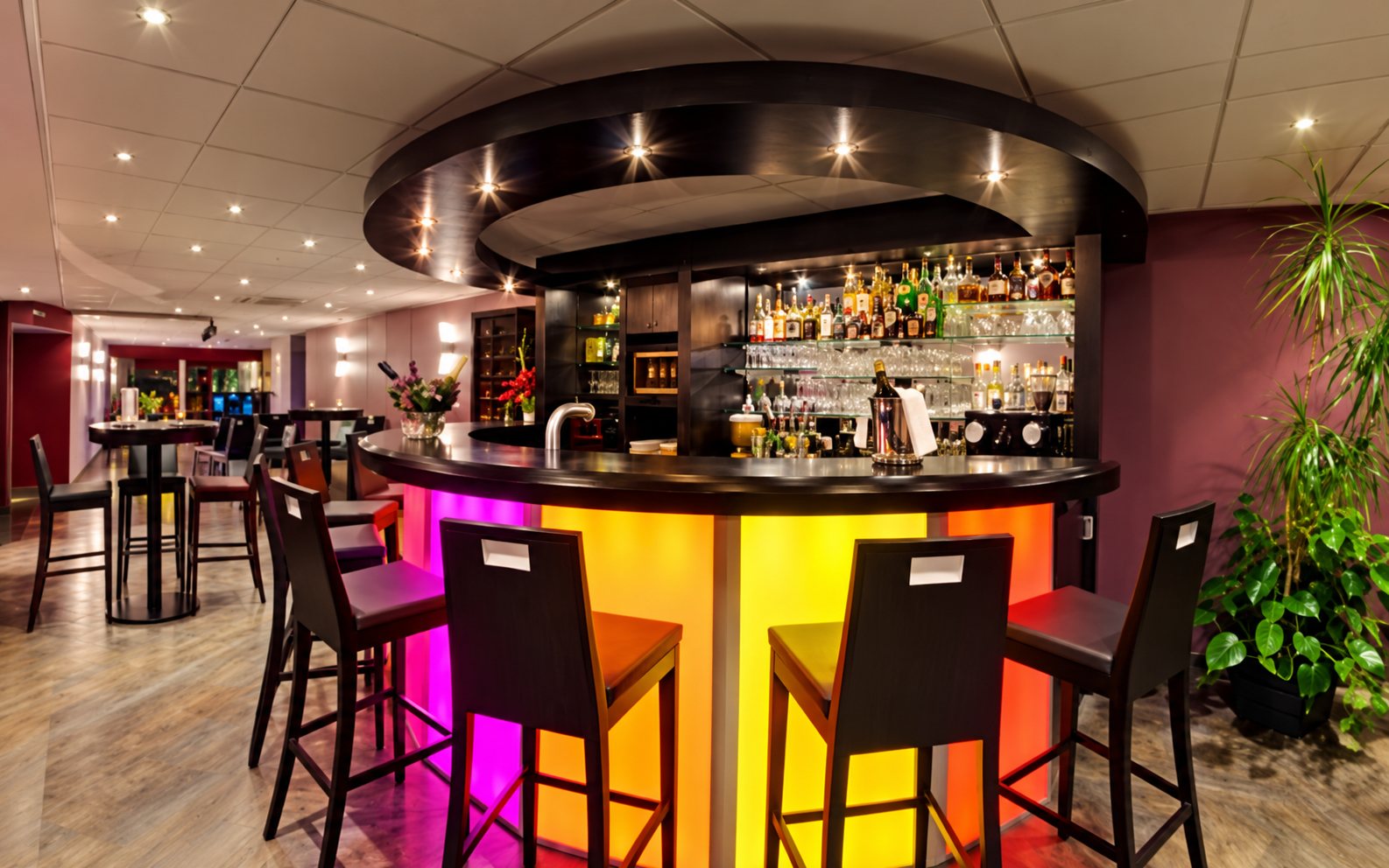 Le bar du hall : comptoir circulaire éclairé en jaune et rose, entouré de tabourets noirs et d’étagères de bouteilles.