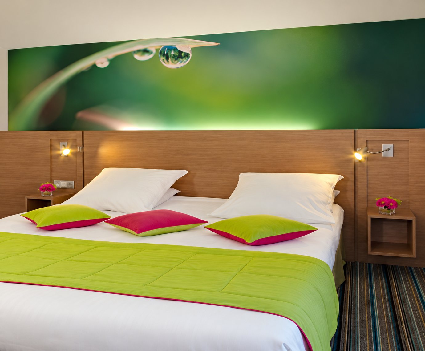 Chambre lumineuse avec un grand lit blanc, un chemin de lit vert anis, des coussins colorés et une grande image de goutte d’eau sur une feuille au-dessus de la tête de lit.