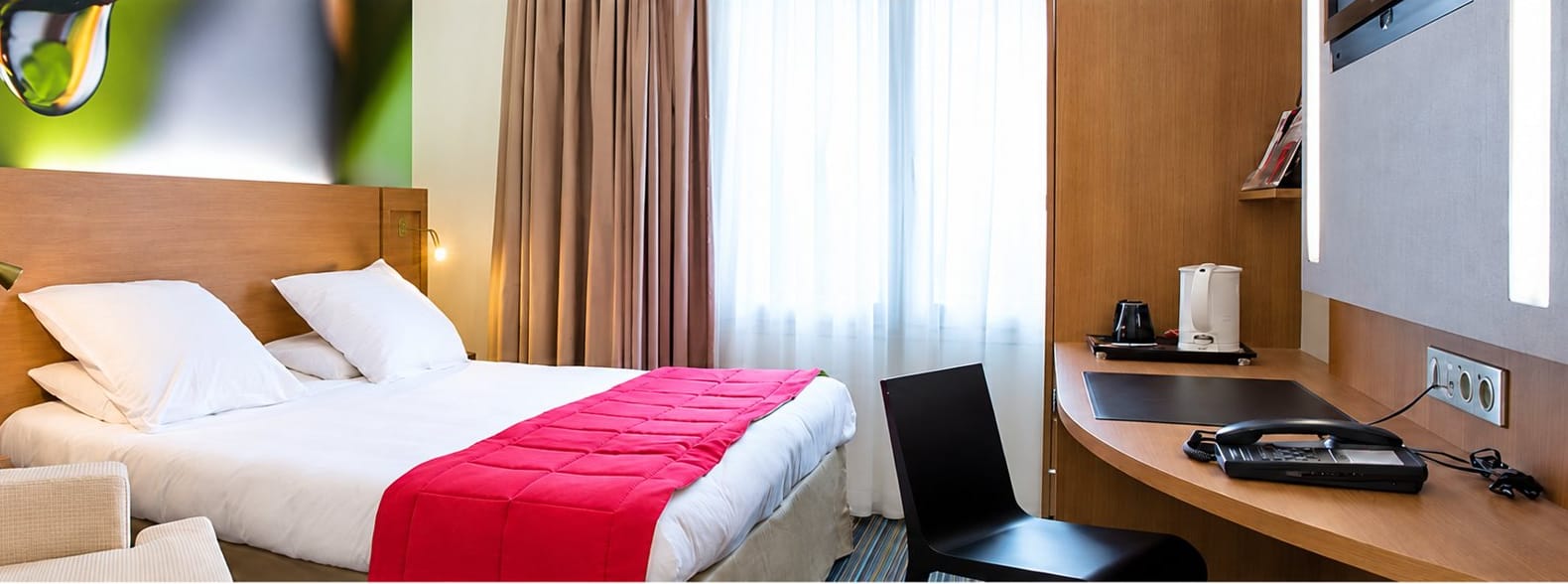 Chambre moderne avec un lit double couvert d’un plaid rouge, un fauteuil beige et un bureau avec téléphone et bouilloire.