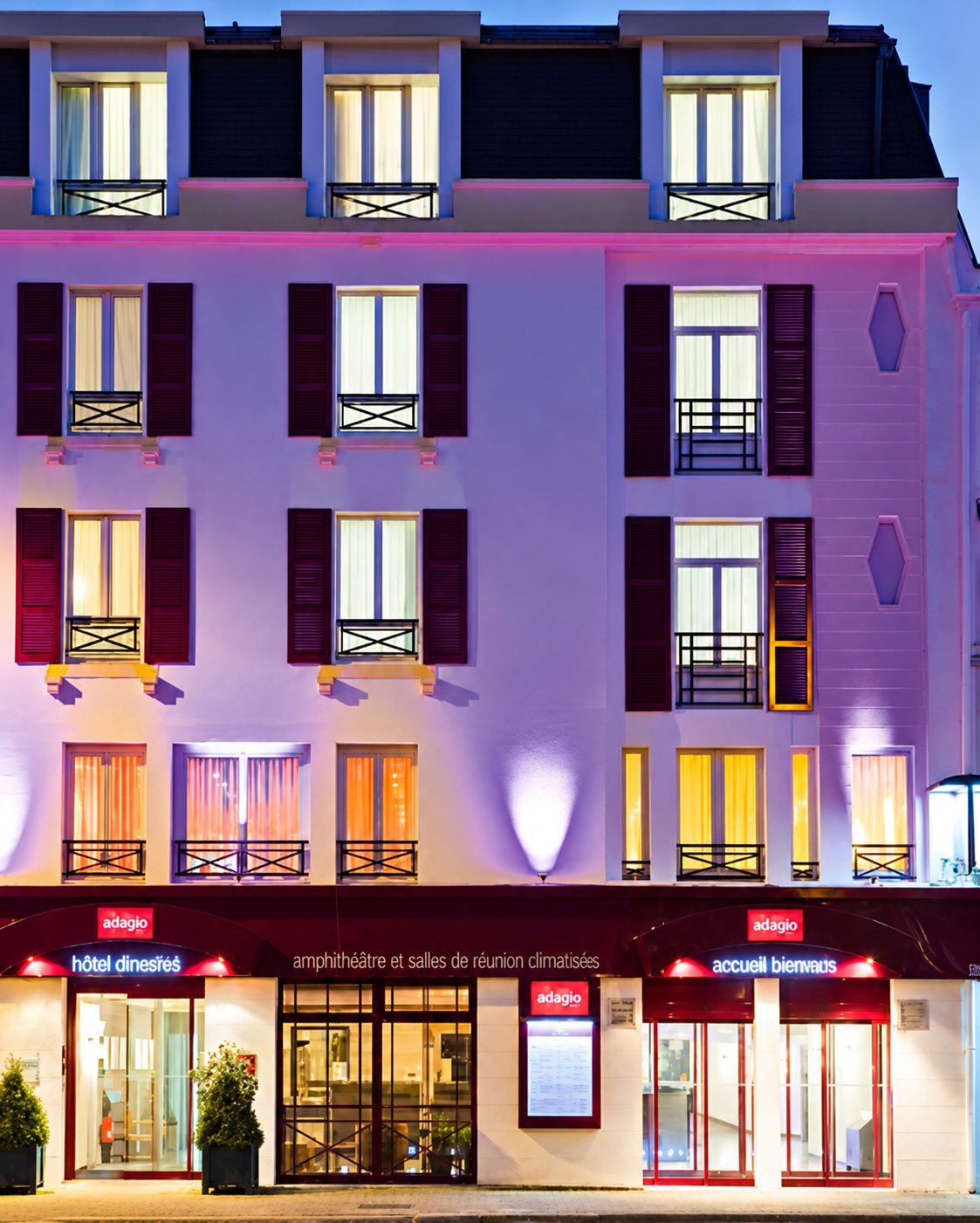 Façade de l’hôtel Mercure Quimper éclairée en violet et rose à la tombée de la nuit, avec les enseignes rouges du rez-de-chaussée.