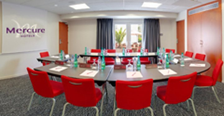 Salle de réunion avec des tables disposées en U, des chaises rouges et des bouteilles d’eau sur les tables.