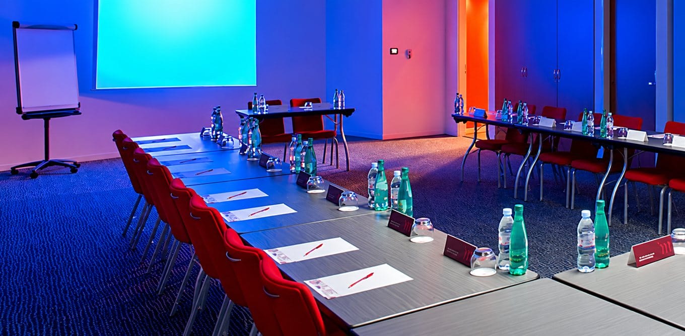 Salle de réunion éclairée en bleu et rose, avec des tables disposées en U, des bouteilles d’eau, des chevalets nominatifs et des chaises rouges.