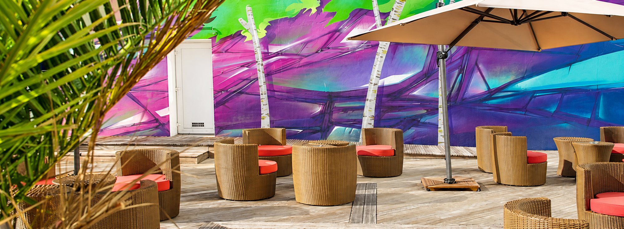 Terrasse en bois avec des fauteuils en rotin à coussins rouges sous un grand parasol, devant une fresque murale violette et verte.