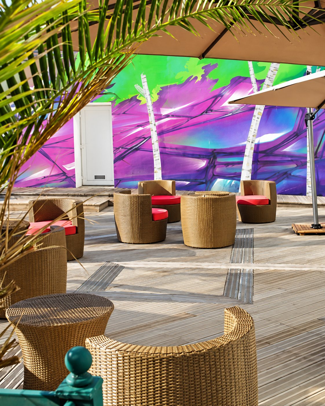 Terrasse en bois meublée de fauteuils et tables en rotin à coussins rouges, devant un mur orné d’une fresque verte et violette, sous des parasols et des palmes.